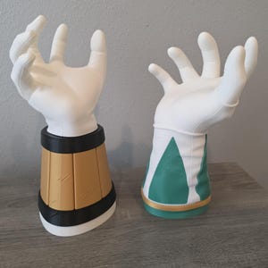 May include: Two white hand-shaped display stands. The left stand features a gold and black cylindrical base with etched lines. The right stand features a white and green cuff design with a gold trim at the bottom. Both hands are in an open, grasping pose.
