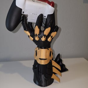 May include: A black and gold gauntlet-shaped stand holding a white and black Xbox controller. The stand features articulated fingers and a forearm piece with gold accents. The controller has a white body, black grips, and red top buttons.