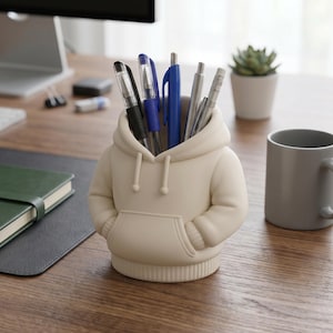 May include: A cream-colored pen holder shaped like a hoodie, filled with pens and pencils. The hoodie-shaped holder has a front pocket and a hood. A gray mug and a small potted plant are in the background.