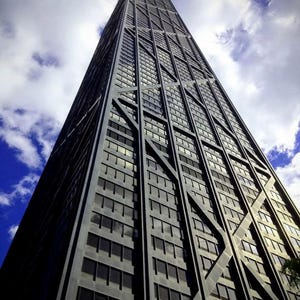 Può includere: Un grattacielo alto e scuro con un caratteristico design esterno a traliccio, visto dal basso contro un cielo parzialmente nuvoloso. La facciata dell'edificio presenta numerose finestre rettangolari, che riflettono il cielo. L'altezza e l'architettura unica della struttura sono enfatizzate.