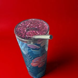 May include: A stainless steel tumbler with a floral design in shades of blue and pink. The lid is filled with pink glitter, and a silver straw is inserted. The tumbler is set against a solid red background.