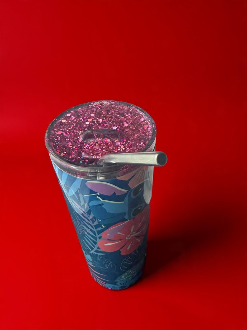 May include: A stainless steel tumbler with a floral design in shades of blue and pink. The lid is filled with pink glitter, and a silver straw is inserted. The tumbler is set against a solid red background.