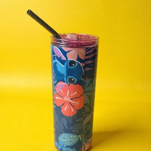 May include: A tall, clear tumbler with a colorful design featuring a cartoon character and tropical flowers. The tumbler has a black straw and is filled with a purple liquid. The background is a solid yellow.
