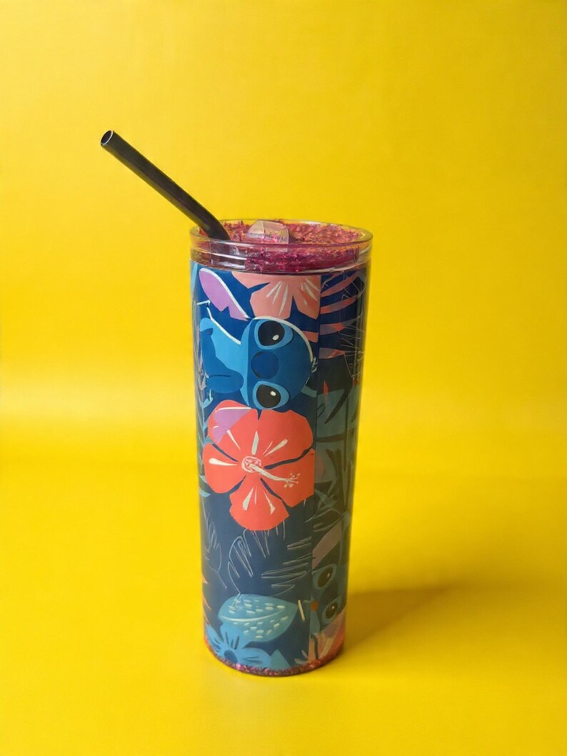 May include: A tall, clear tumbler with a colorful design featuring a cartoon character and tropical flowers. The tumbler has a black straw and is filled with a purple liquid. The background is a solid yellow.