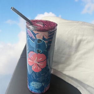 May include: A blue tumbler with a floral and character design, featuring a pink glitter lid and a silver straw. The tumbler has a tropical theme with pink and white flowers and a cartoon character.