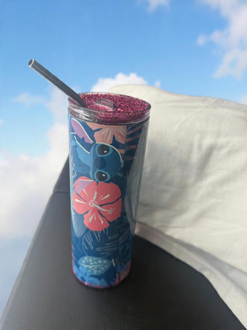 May include: A blue tumbler with a floral and character design, featuring a pink glitter lid and a silver straw. The tumbler has a tropical theme with pink and white flowers and a cartoon character.