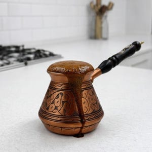 Op de afbeelding: Een koperen cezve, een traditionele Turkse koffiepot, die overloopt van vers gezette koffie. De pot heeft ingewikkelde geëtste ontwerpen en een zwarte houten handgreep. De koffie is een diepbruine kleur en de pot staat op een wit oppervlak.
