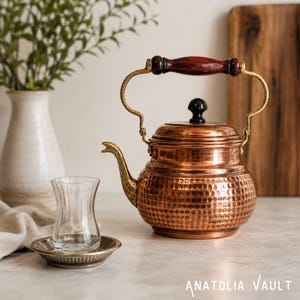 May include: A hammered copper tea kettle with a dark wooden handle, a brass-toned spout, and a black knob on the lid. Beside it sits a clear glass tea cup on a small metallic saucer. The text Anatolia Vault is visible in the bottom right corner.