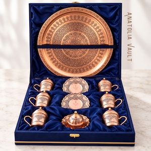 May include: A copper coffee set in a blue velvet-lined box, featuring a large round tray, six cups with lids, and three saucers, all engraved with intricate black geometric patterns. The text ANATOLIA VAULT is printed on the right side of the box.