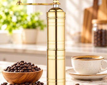 May include: A cylindrical gold-colored manual coffee grinder with a top-mounted crank handle. The grinder features a polished metallic finish with horizontal grooved detailing along the body and a small knob on the end of the handle.