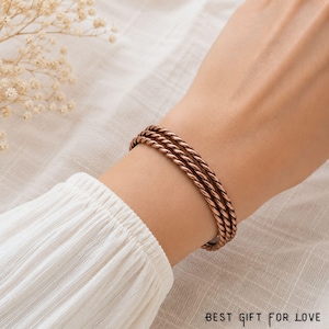 May include: A copper-toned bracelet featuring three intertwined twisted metal strands that form a continuous rope-like design. The bracelet is worn on a wrist, and the text BEST GIFT FOR LOVE is printed in the bottom right corner of the image.