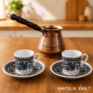 Peut inclure: Une cafetière en cuivre avec une poignée noire et des motifs gravés, accompagnée de deux tasses en céramique blanche et soucoupes assorties avec un motif floral bleu, rouge et vert. Le texte Anatolia Vault est visible en bas à droite.