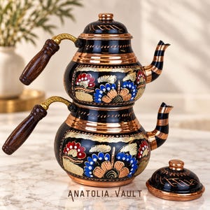 Op de afbeelding: Een tweekoppige koperen theepotset met een zwarte emaille afwerking en handgeschilderde bloemmotieven in rood, blauw en goud. De set bevat twee gestapelde potten met donkere houten handvatten, goudkleurige accenten en een bijpassend los deksel.