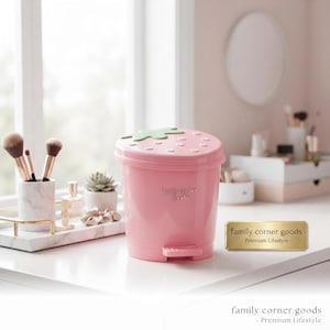 May include: A pink plastic trash can with a strawberry-shaped lid featuring green leaves and white dots. The front of the bin is embossed with the text family corner goods. It includes a foot pedal at the base for opening the lid.