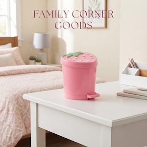 Op de afbeelding: Een roze cilindrische afvalemmer met een aardbeivormig deksel met een groen steeltje en kleine verhoogde stippen. De afvalemmer is voorzien van een voetpedaal om het deksel te openen. De tekst FAMILY CORNER GOODS is zichtbaar boven in de afbeelding.