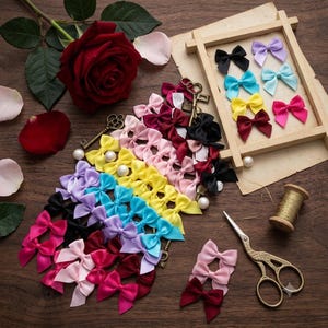 Puede incluir: Una colección de pequeños lazos de cinta de satén en varios colores, incluyendo rojo, rosa, amarillo, azul, morado y negro, dispuestos junto a un marco de madera, dos llaves de metal de estilo antiguo, un carrete de hilo dorado y tijeras.