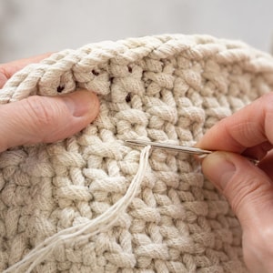May include: A thick, off-white crocheted textile made from heavy cord, featuring a dense, textured stitch pattern. A thin, metallic sewing needle is inserted into the fabric, pulling a fine, matching off-white thread through the loops.