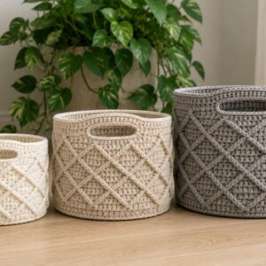 May include: Three round, crocheted storage baskets in graduated sizes, featuring a textured diamond lattice pattern on the exterior and integrated oval handles at the top rim. The two smaller baskets are cream-colored, while the largest is gray.