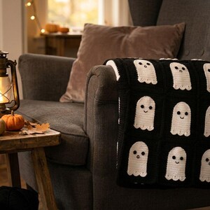 May include: A black and white crochet blanket featuring a repeating pattern of white ghosts with dark eyes and mouths on a black background. The blanket is draped over a dark gray armchair, showcasing a grid of individual square motifs.