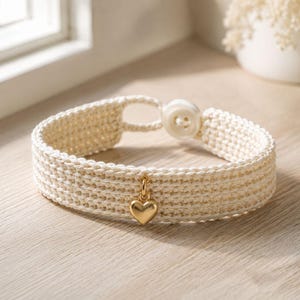 Puede incluir: Una pulsera de ganchillo de color crema hecha de hilo grueso, con un pequeño dije de corazón dorado pulido que cuelga del centro y un cierre de botón blanco único en el extremo de la banda, creando un accesorio de muñeca sencillo y texturizado.
