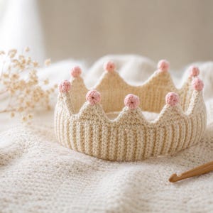 Può includere: Una corona all'uncinetto color crema caratterizzata da una fascia a coste e punte sormontate da piccole sfere all'uncinetto rosa chiaro. La corona è realizzata con un motivo a maglia testurizzato e presenta un design uniforme e coerente.