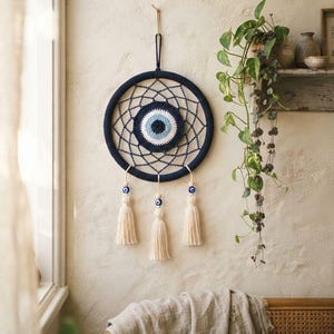 Könnte beinhalten: Ein kreisförmiger Wandbehang mit einem dunkelblau umhäkelten Ring und einem zentralen Schutzaugen-Motiv in Weiß, Hellblau und Dunkelblau. Drei hängende Schnüre sind unten befestigt, jede mit einer kleinen Schutzaugen-Perle und einer cremefarbenen Quaste.