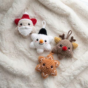 May include: Four star-shaped crochet ornaments featuring holiday characters: a Santa Claus with a red hat, a snowman with a black hat and orange nose, a brown reindeer with antlers and a red nose, and a tan gingerbread star with white icing details.