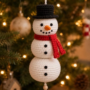 May include: A crocheted snowman ornament featuring a white body, a black top hat, a bright red scarf, and an orange carrot nose. The snowman has four black buttons on its torso and a small wooden bead hanging from the bottom on a white string.