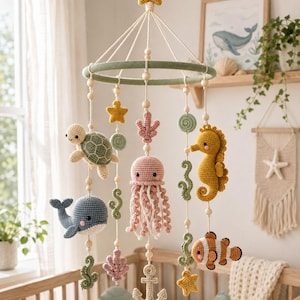 May include: A nursery mobile featuring crocheted sea creatures including a turtle, whale, jellyfish, seahorse, and clownfish, along with stars, coral, and an anchor. The items are suspended from a green ring with wooden beads on white strings.