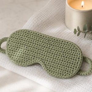 May include: A sage green crochet sleep mask featuring a textured, hand-stitched pattern with a rounded, contoured shape and two circular side loops for securing the mask in place. The item is crafted from yarn with a consistent, dense stitch.