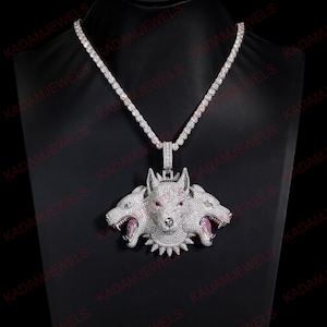 May include: A silver-toned necklace featuring a large, three-headed wolf pendant encrusted with clear, white, and red stones. The pendant hangs from a matching tennis-style chain, with the text KADAMJEWELS repeated across the dark background.