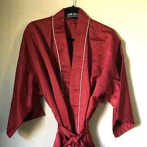 May include: A burgundy satin robe with white trim and a matching tie. The robe is hanging on a black hanger. The text "THE BLU" is visible on the hanger.