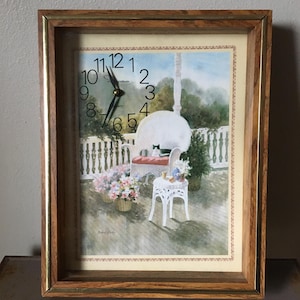 May include: A framed clock with a painted scene of a porch with a white wicker chair, flowers, and a cat. The clock face has black hands and Roman numerals. The frame is brown with a gold inner edge.