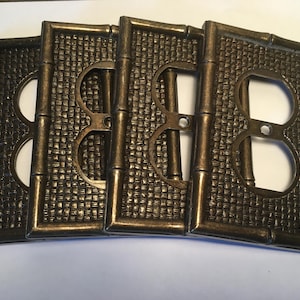 May include: A set of four rectangular bronze-colored metal wall plates for electrical outlets. Each plate features a textured woven pattern in the center and a raised border designed to resemble bamboo stalks with a central screw hole.