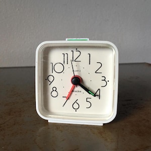 Puede incluir: Un reloj despertador de cuarzo cuadrado blanco con una esfera color crema. El reloj tiene números negros, un segundero rojo, un minutero negro y un indicador de alarma verde. La marca "Westclox" está impresa en la esfera.