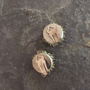 May include: A pair of round, silver clip-on earrings with a light blue and gold beaded design.