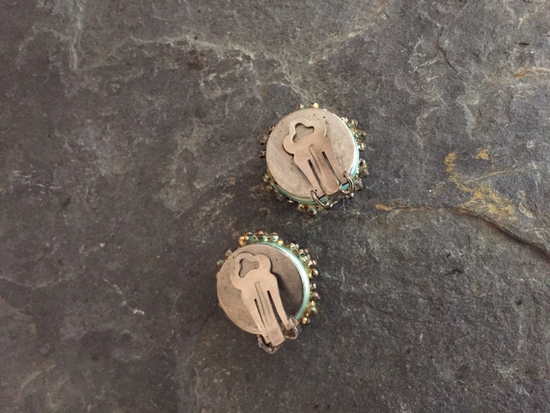 May include: A pair of round, silver clip-on earrings with a light blue and gold beaded design.