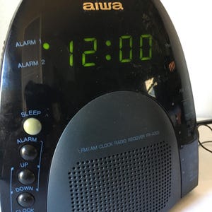 May include: A black Aiwa FM/AM clock radio receiver model FR-A305 with a digital display showing 12:00 in green. The device features a rounded top, a front-facing speaker grille, and a vertical column of circular control buttons on the left side.