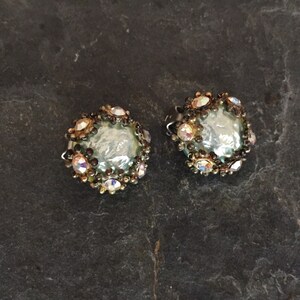 May include: A pair of silver tone clip-on earrings with a round, iridescent, light blue stone surrounded by clear and black rhinestones.