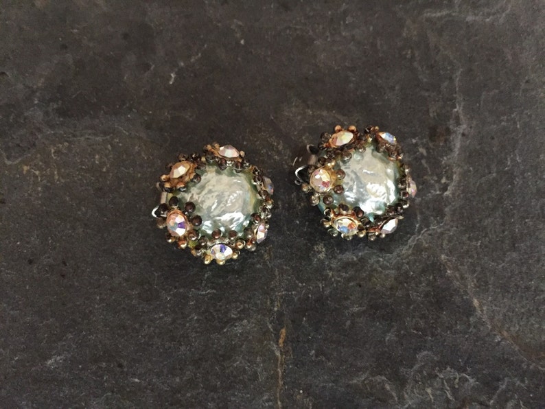 May include: A pair of silver tone clip-on earrings with a round, iridescent, light blue stone surrounded by clear and black rhinestones.