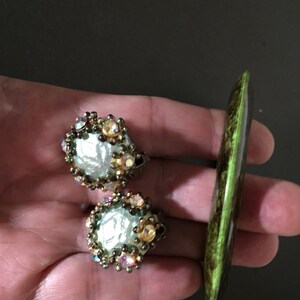 May include: A pair of gold tone earrings with a large, iridescent, white pearl-like center surrounded by clear and iridescent rhinestones. The earrings are next to a green and brown, long, oval-shaped, decorative object.