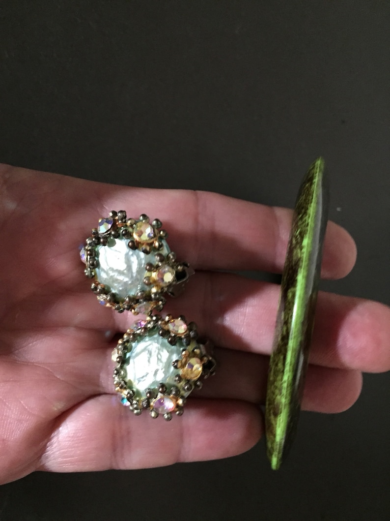 May include: A pair of gold tone earrings with a large, iridescent, white pearl-like center surrounded by clear and iridescent rhinestones. The earrings are next to a green and brown, long, oval-shaped, decorative object.