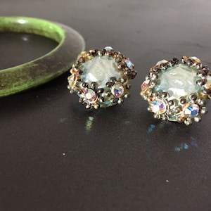 May include: A pair of iridescent blue glass earrings with a silver and gold metal setting. The earrings are surrounded by small, clear rhinestones.