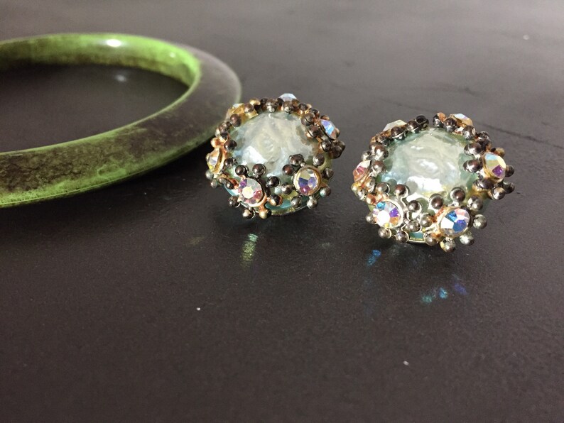 May include: A pair of iridescent blue glass earrings with a silver and gold metal setting. The earrings are surrounded by small, clear rhinestones.