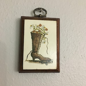 May include: A framed illustration of a brown boot with white laces and flowers growing out of the top. The boot is on a wooden base and the frame has a metal hanger.