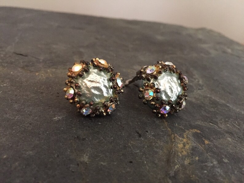 May include: A pair of silver and gold tone earrings with iridescent rhinestones. The earrings feature a round, textured center piece.