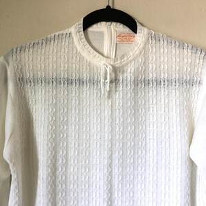 May include: A white long-sleeved shirt featuring a high neckline with a small tie detail and a vertical textured knit pattern. The garment includes a back zipper closure and a visible interior label at the neck area.