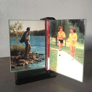 May include: A black plastic rotating photo display stand featuring two clear acrylic panels. The left panel shows a person standing by a lake, while the right panel displays two people jogging on a path. The stand has a central red accent piece.