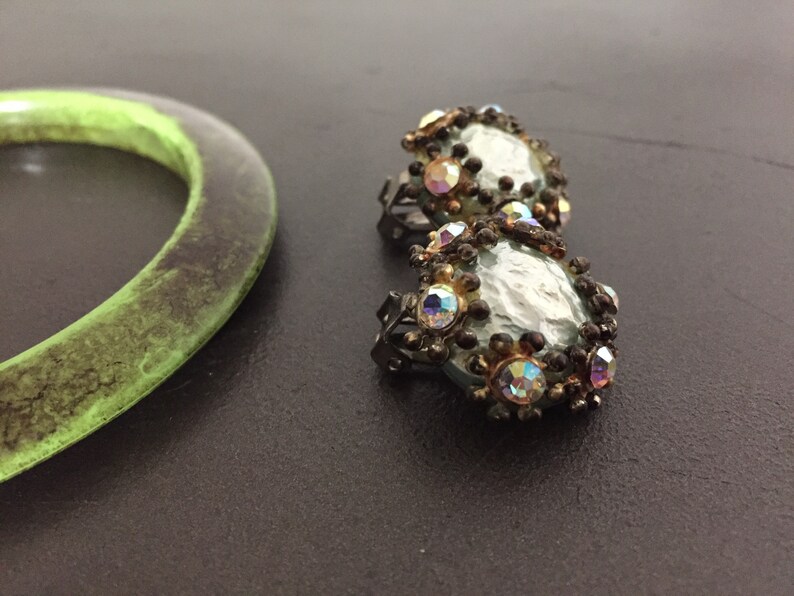 May include: A pair of silver tone earrings with iridescent rhinestones and a light blue center. The earrings are on a dark gray surface.