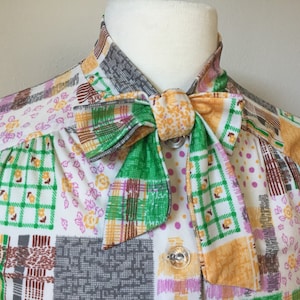 May include: A white blouse with a patchwork design featuring green, brown, orange, and pink squares. The blouse has a large bow tie made from the same fabric.